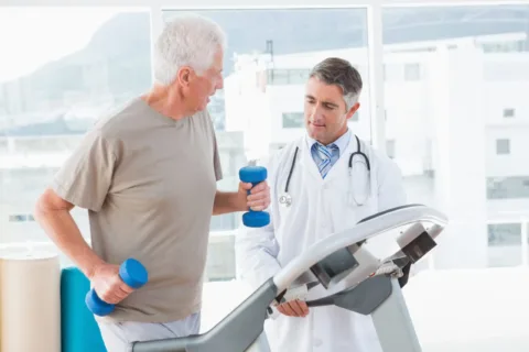 elderly man jogging on a treadmill holding weights while a doctor stands next to him